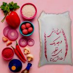 بسته برنج هاشمی درجه یک با بسته‌بندی سنتی، همراه با چیدمانی از مواد غذایی تازه مانند فلفل قرمز، پیاز بنفش، سیر، ادویه، روغن، گوشت و سبزیجات روی زمینه صورتی